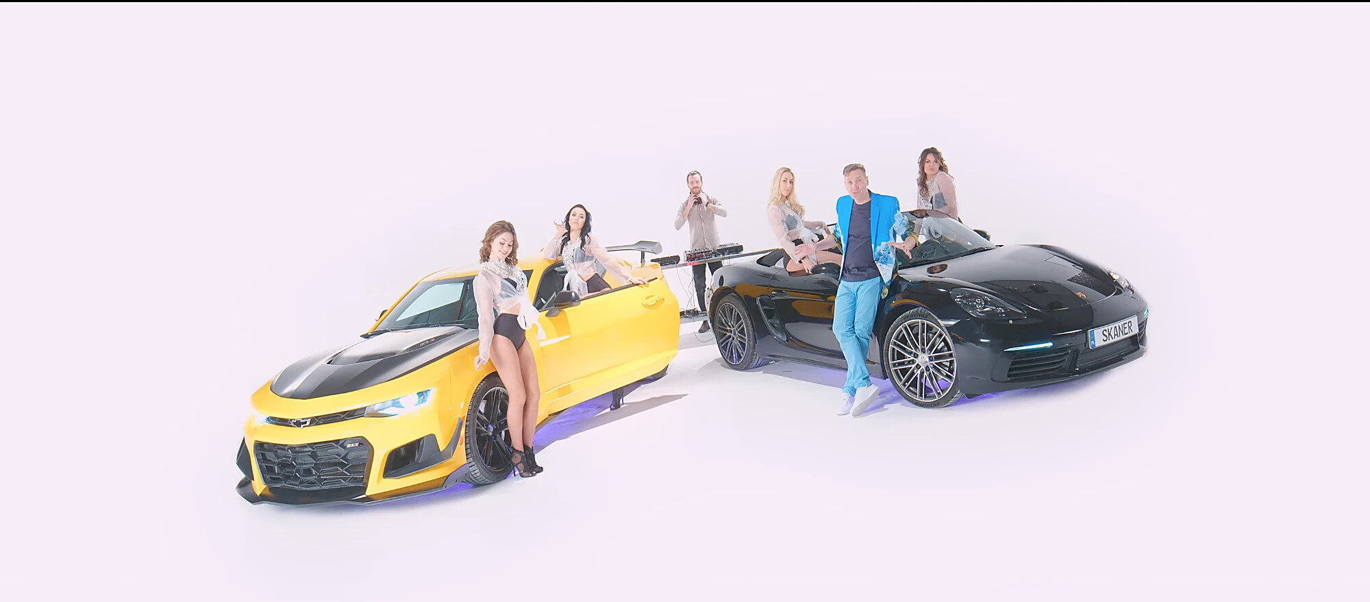 Zespół Skaner w studiu: Robert Sasinowski w błękitnej marynarce opiera się o czarne Porsche z tablicą SKANER, obok żółte Chevrolet Camaro, tancerki i DJ