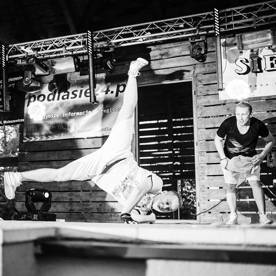 Czarno-białe zdjęcie: tancerz w akrobatycznej figurze na scenie festynu, obok Robert Sasinowski